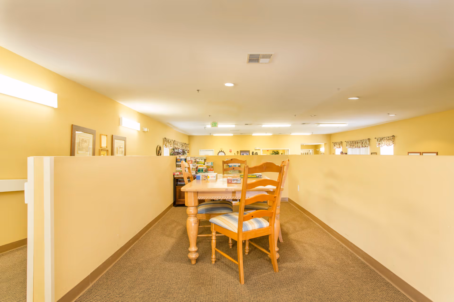 A well-lit common area in a senior living facility with a wooden table and four wooden chairs with striped cushions. The table has several board games stacked on it. The walls are painted yellow and decorated with framed pictures. The carpet is brown, and there are windows with floral valances in the background.