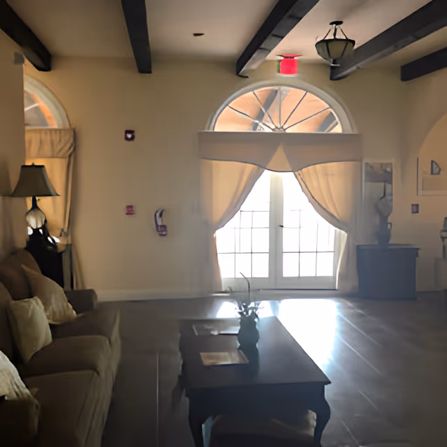 A cozy living room area with a beige sofa adorned with cushions, a wooden coffee table with a vase and some papers on it, and a large arched window with sheer curtains allowing natural light to fill the room. The ceiling has exposed dark wooden beams and a hanging light fixture.