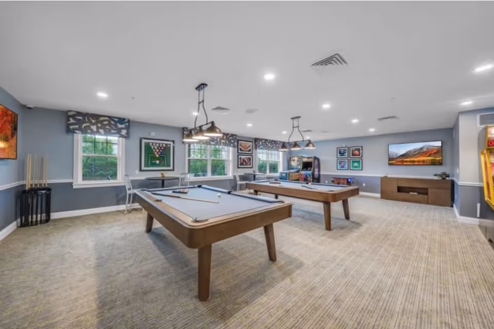 Bright recreational game room with two pool tables, hanging lights, a wall-mounted TV and arcade machines.