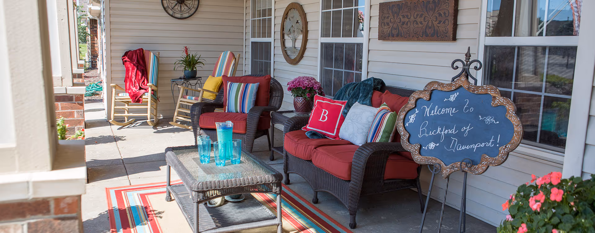 Outdoor patio area with wicker furniture including a loveseat and chairs with red cushions and colorful pillows. A glass-top coffee table with blue glasses sits on a striped rug. A decorative chalkboard sign reads 'Welcome to Bickford of Davenport!' There are potted plants and wall decorations on the siding of the building.