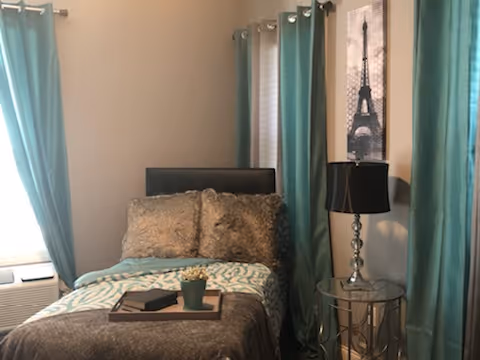 A cozy bedroom with a single bed featuring a brown and teal patterned comforter and two large pillows. Next to the bed is a round glass-top side table with a decorative lamp. The room has teal curtains on two windows, and a framed picture of the Eiffel Tower hangs on the wall.