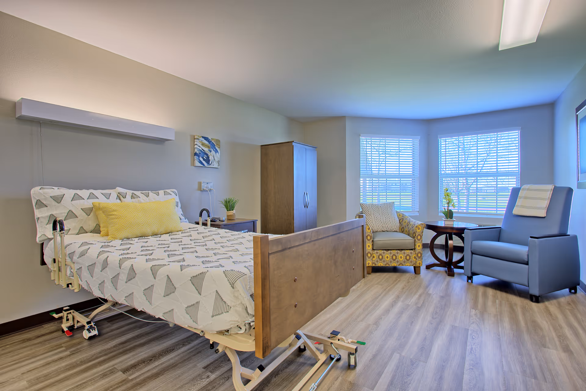 A bright and clean bedroom in a senior living facility featuring a hospital-style bed with patterned bedding and a yellow pillow. The room has wood flooring, a wooden wardrobe, a nightstand with a small plant, two armchairs near a round wooden table by large windows with blinds, and a piece of abstract wall art above the bed.