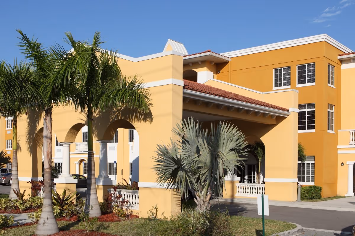 Exterior view of a yellow and orange multi-story assisted living facility building with a covered entrance supported by columns, surrounded by palm trees and landscaping under a clear blue sky.