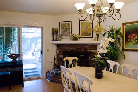 A dining room with a rectangular table surrounded by six white chairs. A chandelier with multiple lights hangs above the table. There is a brick fireplace with a white mantle decorated with framed pictures, candles, and a flower arrangement. To the left, a sliding glass door leads outside, and to the right, there is a large green plant and a framed painting of fruit on the wall.