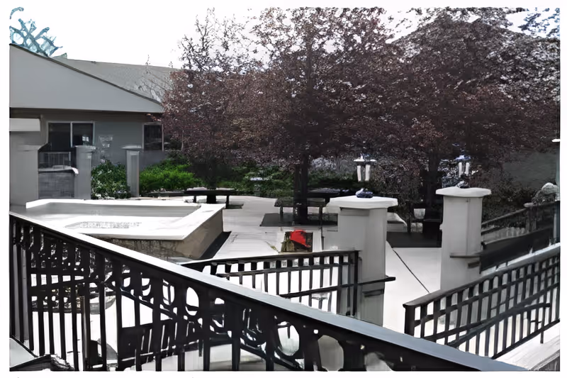 Outdoor courtyard with iron railings, paved walkways, benches, a raised fountain, and trees.