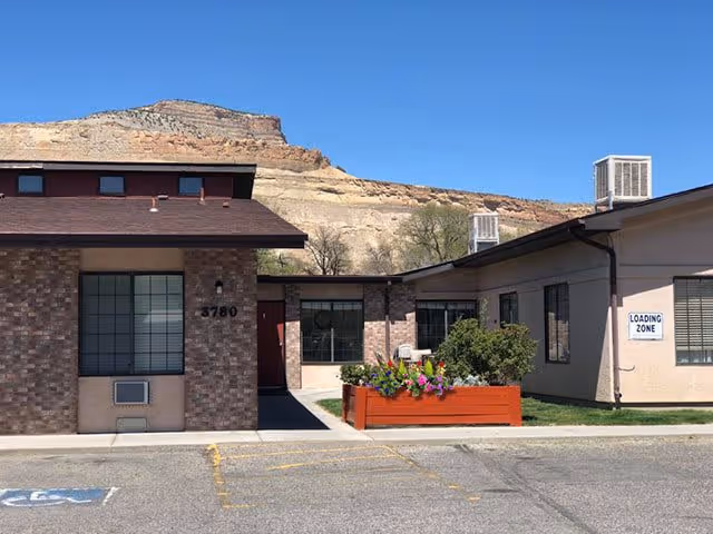 Exterior view of a single-story senior living facility building with brick and beige walls, windows, and a red flower planter in front. There is a mountain in the background under a clear blue sky. A sign on the building reads 'LOADING ZONE' and the building number '3780' is visible.