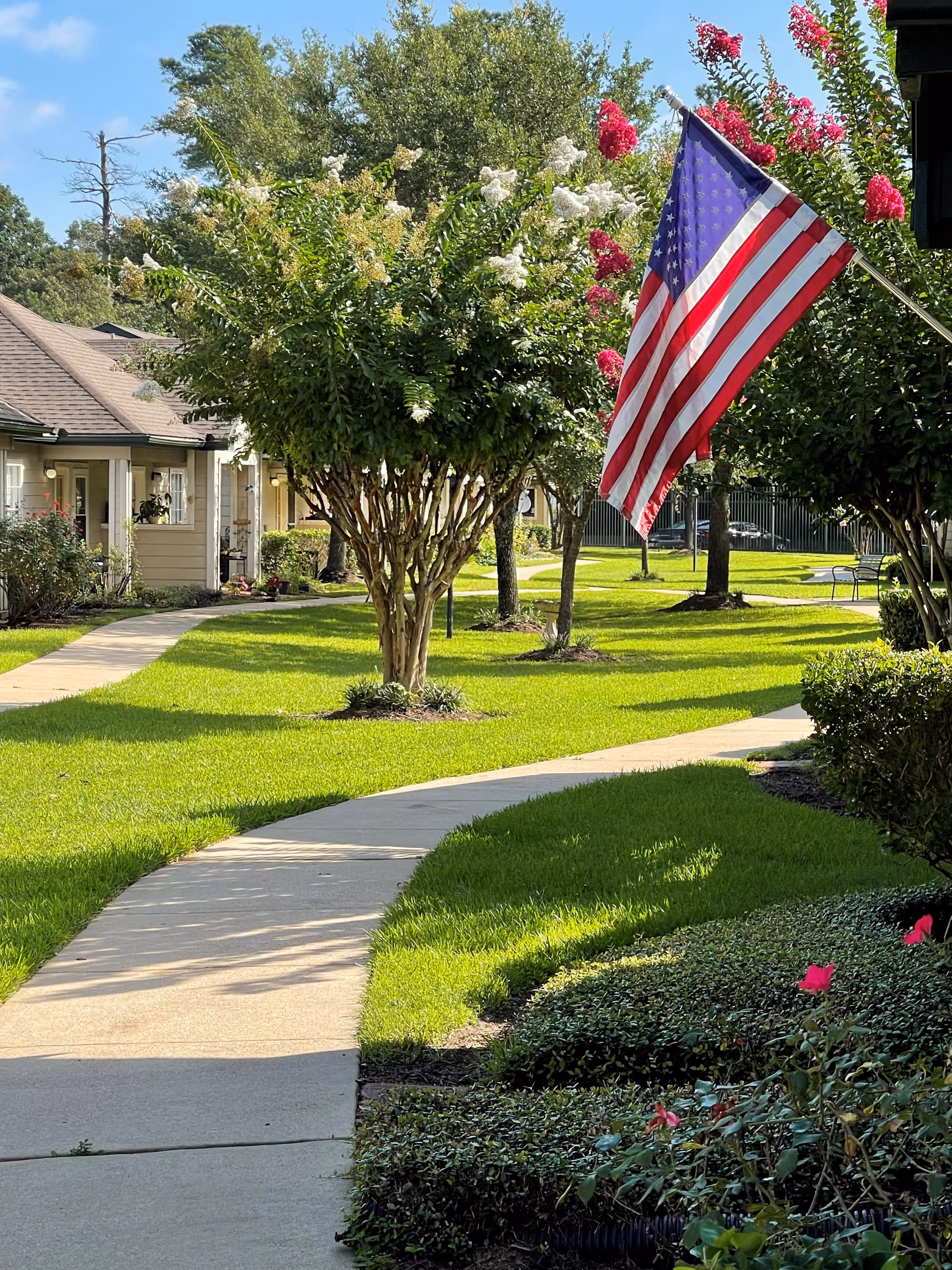A sunny outdoor pathway winding through a well-maintained garden area with green grass, flowering bushes, and trees. An American flag is prominently displayed on a pole attached to a building on the right side. Residential-style buildings with porches are visible on the left side of the pathway.