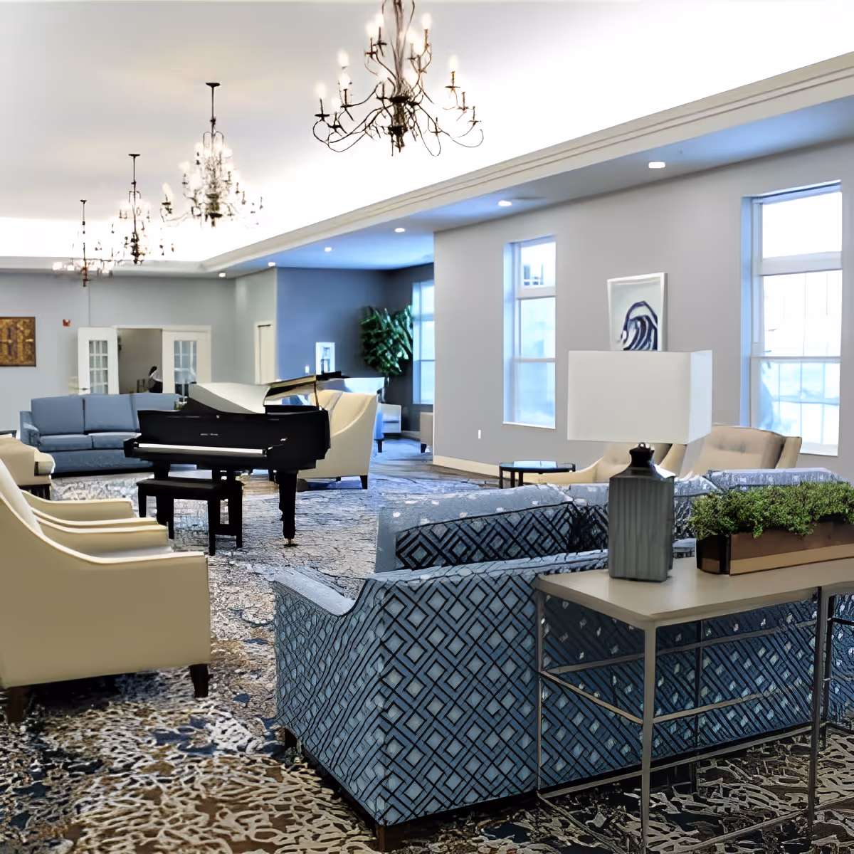 A spacious senior living facility common area with multiple seating arrangements including blue patterned sofas, beige armchairs, and a black grand piano in the center. The room features large windows allowing natural light, decorative chandeliers hanging from the ceiling, and a side table with a lamp and a planter.