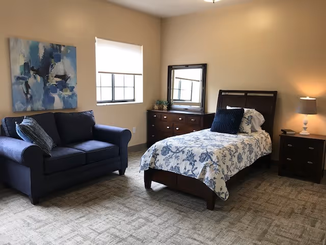A bedroom with a single bed covered in a white and blue floral bedspread, a dark wooden headboard, and a navy blue pillow. Next to the bed is a dark wooden nightstand with a white lamp. Across from the bed is a dark wooden dresser with a mirror and a small plant. To the left of the dresser is a dark blue loveseat with two pillows. A window with a white blind is above the loveseat, and a blue abstract painting hangs on the wall above it. The room has beige walls and carpeted flooring.