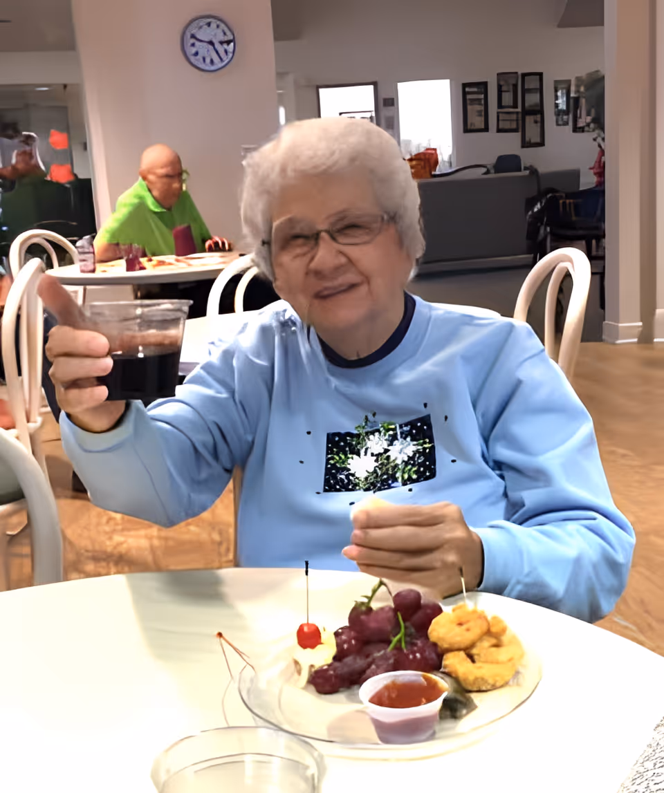 An elderly woman with white hair and glasses is sitting at a table in a dining area, holding up a glass of dark beverage and smiling. On the table in front of her is a plate with grapes, a cherry, some fried snacks, and a small cup of dipping sauce. In the background, another elderly person is seated at a different table, and the room has light-colored walls and wooden flooring.