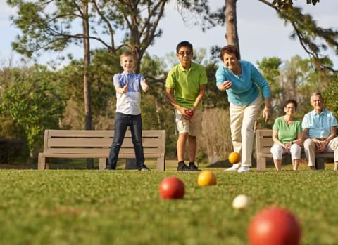 Two children and an elderly woman playing bocce ball on a grassy lawn with trees in the background, while two elderly people sit on a bench watching.