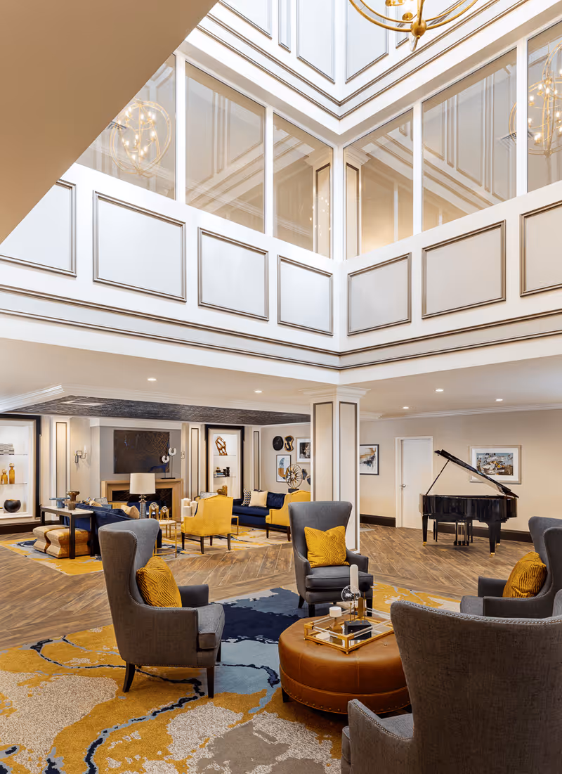 Spacious and elegant living room area with high ceilings and large windows. The room features multiple seating arrangements including gray armchairs with yellow cushions, yellow armchairs, and blue sofas. A round leather ottoman with decorative items is in the center. The floor has a patterned carpet with blue, yellow, and beige tones. A black grand piano is positioned against one wall, and the walls are decorated with framed artwork and shelves with decorative objects. The room is well-lit with recessed lighting and a large chandelier hanging from the ceiling.
