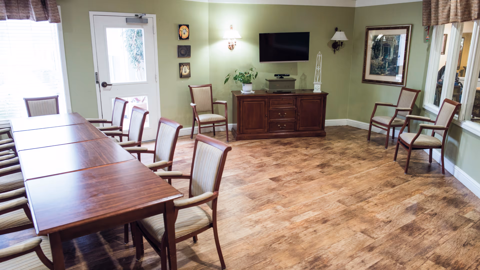 A room with a long wooden table surrounded by chairs with cushioned seats and backs. The walls are painted light green, and there is a wooden cabinet with a flat-screen TV mounted above it. The room has wood flooring, wall sconces, framed artwork, and windows with brown valances.