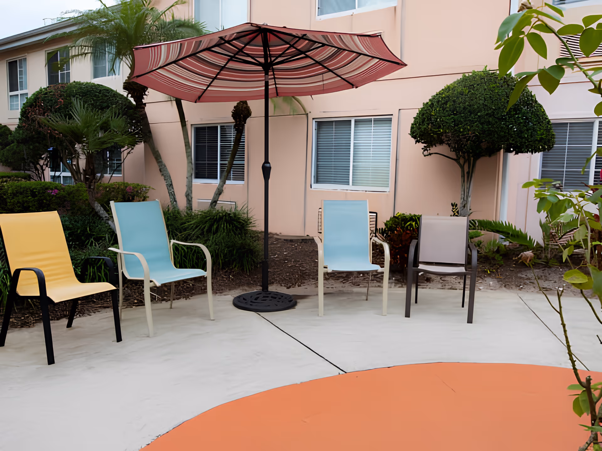 Outdoor courtyard with colorful chairs and a striped patio umbrella in front of a peach-colored building.