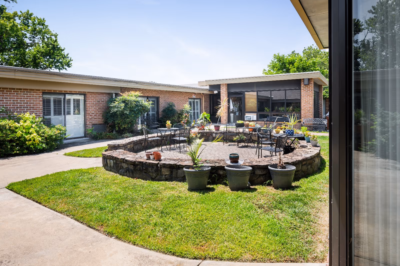 Outdoor courtyard area at Ocean Grove Post Acute featuring a circular stone seating area with metal chairs and tables, surrounded by potted plants and greenery. The courtyard is enclosed by a single-story brick building with large windows and doors, under a clear blue sky.
