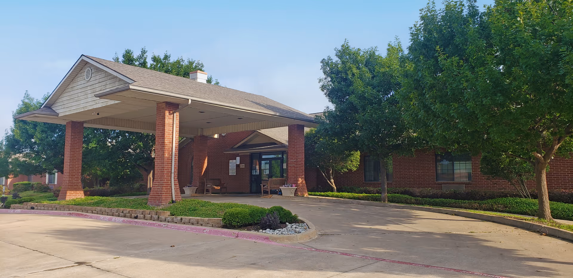 Entrance of a senior living facility with a covered drop-off area supported by brick columns, surrounded by green trees and bushes, and a clear blue sky overhead.
