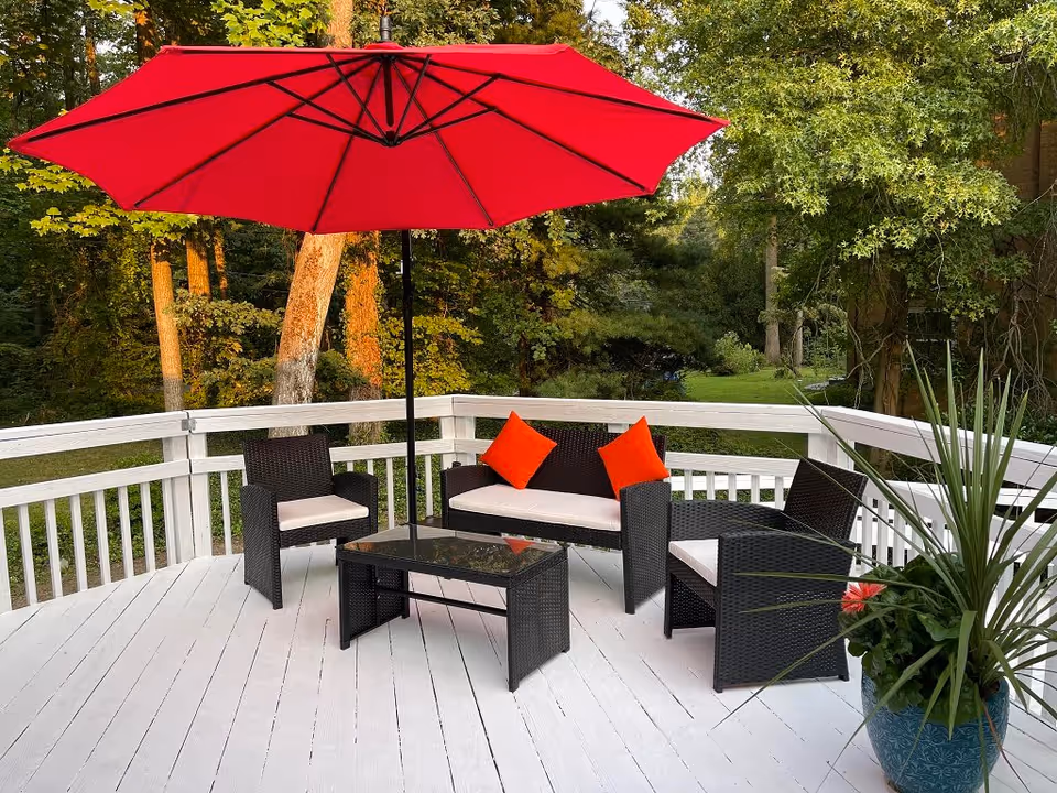 Outdoor patio area with white wooden deck, black wicker furniture including a loveseat with two orange cushions, two armchairs, and a glass-top coffee table. A large red umbrella provides shade, and there is a blue planter with green plants and a red flower on the right side. Trees and greenery are visible in the background.