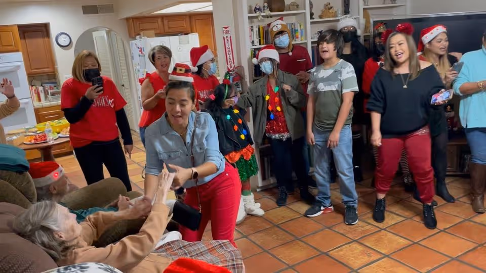 A group of people gathered in a living room decorated for the holidays. Some individuals are wearing Santa hats and festive clothing. One woman in a light blue shirt and red pants is holding hands with an elderly person seated on a couch. Others are standing and appear to be singing or enjoying the event. The room has a tiled floor, wooden cabinets, and a kitchen area visible in the background.