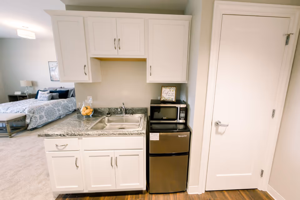 Small kitchenette with white cabinets, a sink and countertop next to a microwave and mini fridge, with a bed visible in the adjacent studio.