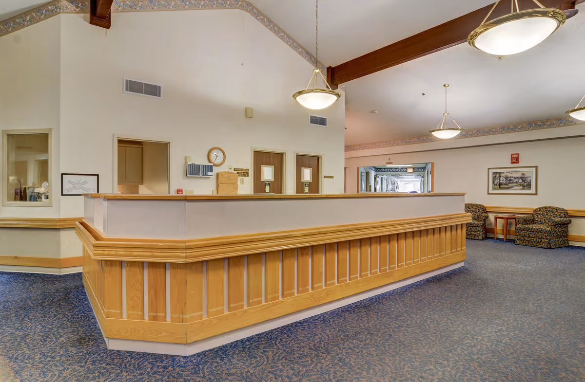 Interior view of a senior living facility reception area with a wooden front desk, blue patterned carpet, beige walls, and ceiling lights. There are two patterned armchairs and a small table in the background near a framed picture on the wall.