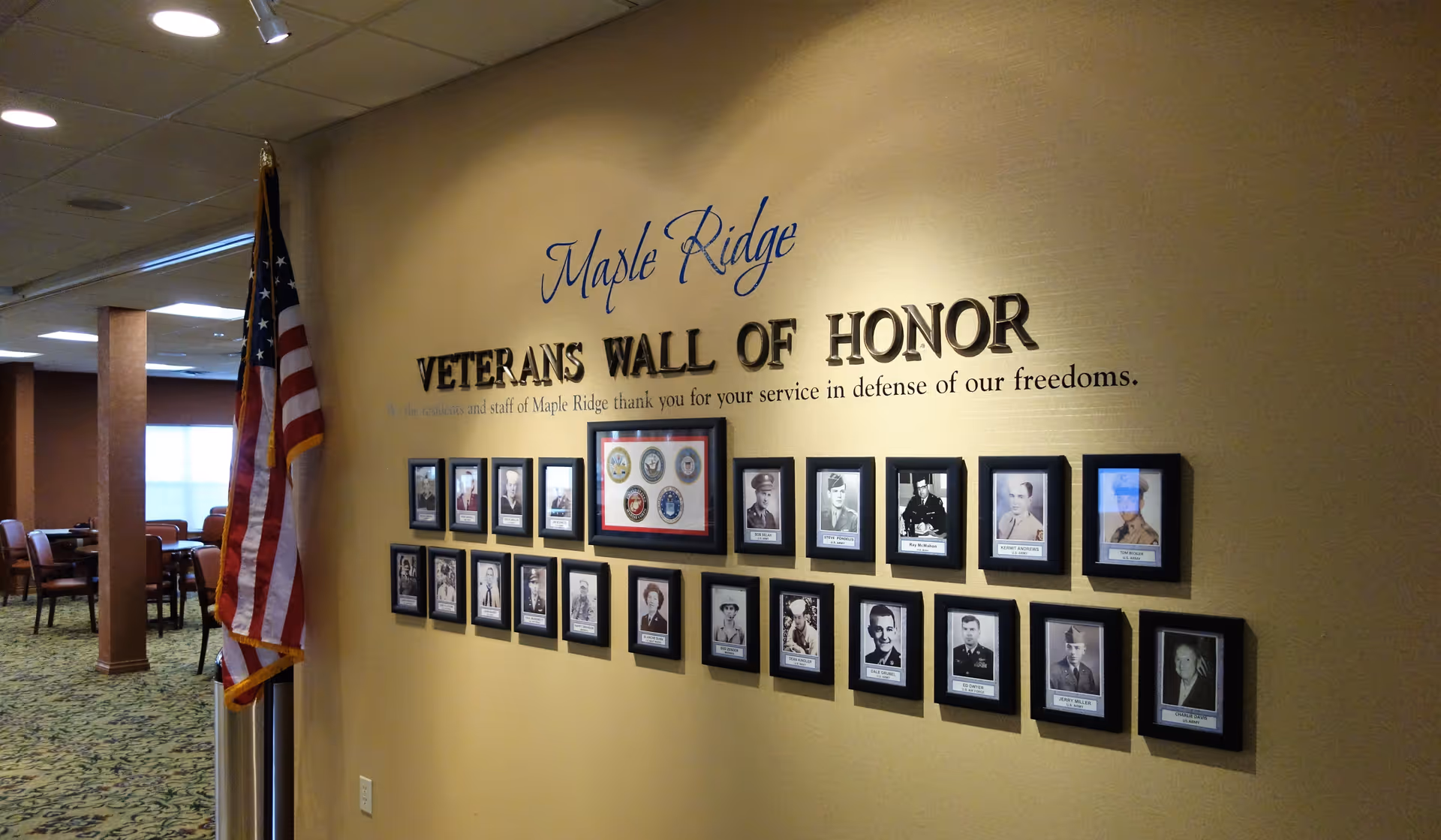 Interior wall of Maple Ridge Retirement Community featuring a Veterans Wall of Honor with framed photos of veterans and an American flag standing beside the wall. The wall has a message thanking veterans for their service in defense of freedoms.
