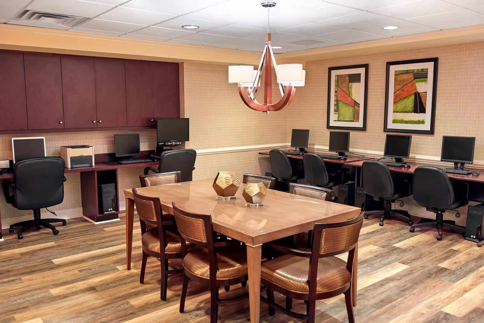 A computer room with a large wooden table surrounded by six chairs in the center. Along the walls, there are desks with multiple desktop computers and office chairs. The room has wood-patterned flooring, beige walls with a chair rail, two abstract framed paintings, and a modern chandelier hanging above the table.
