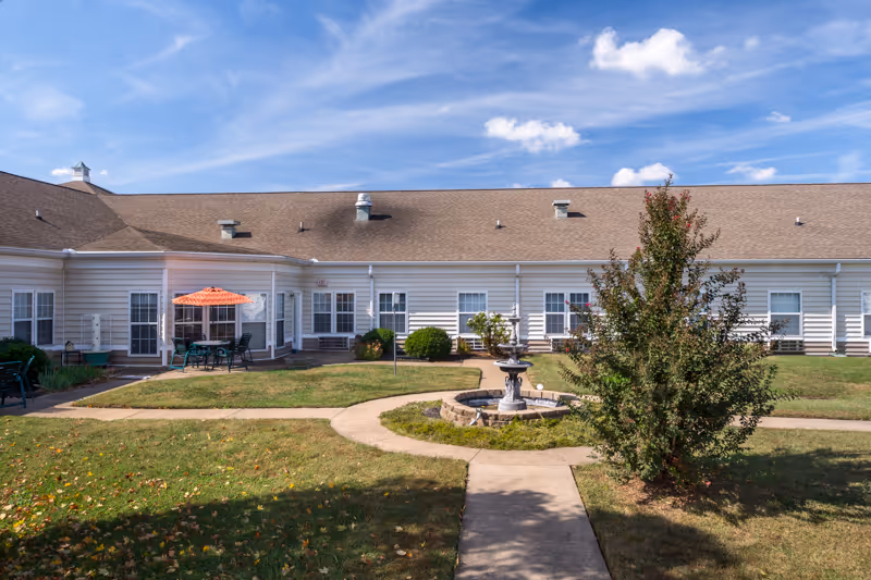 Outdoor courtyard area of a senior living facility with a central fountain, paved walkways, green grass, a tree, and a seating area with a table and orange umbrella. The building has white siding and multiple windows under a brown shingled roof, with a clear blue sky above.