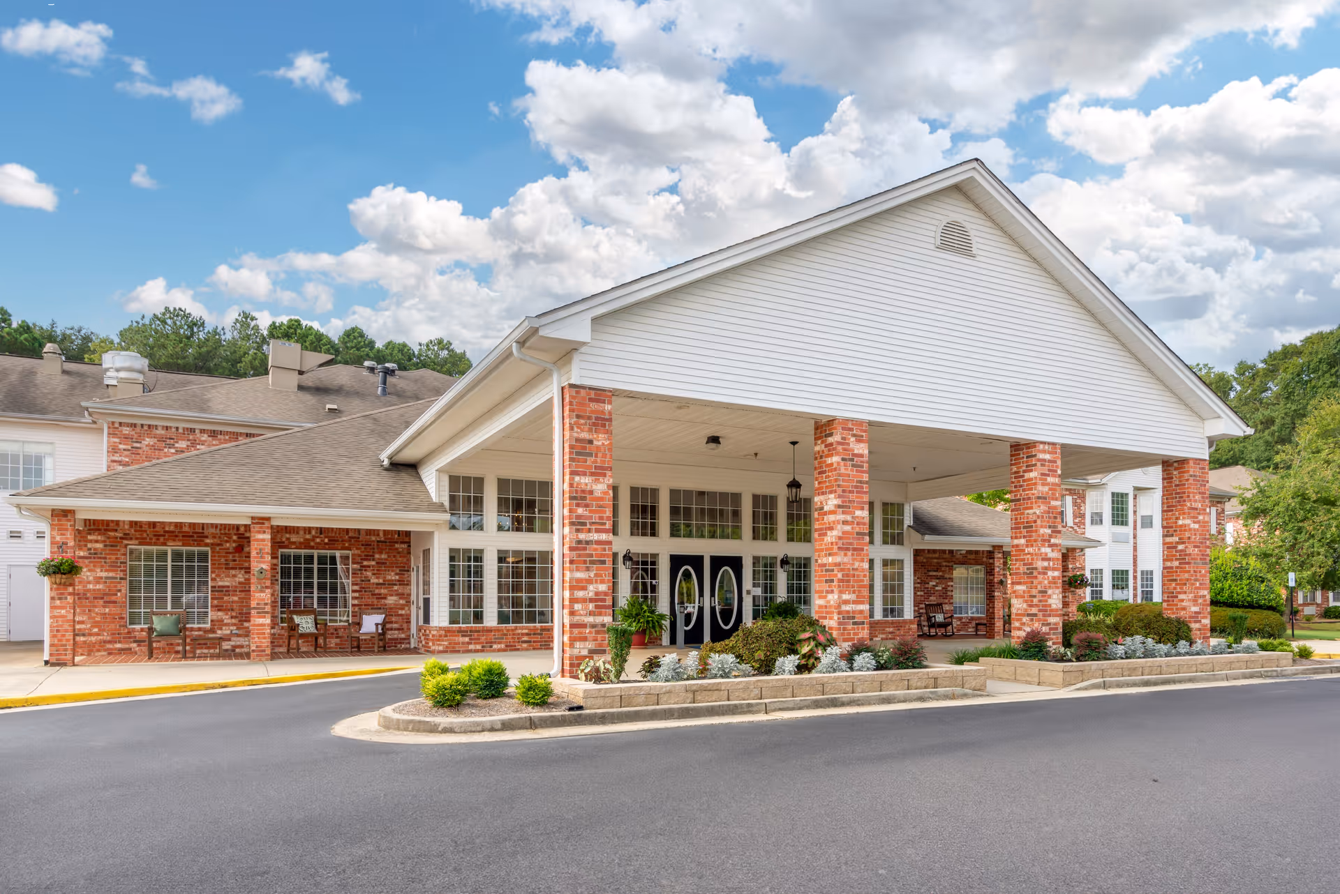 Front exterior view of a senior living facility with a covered entrance supported by brick columns, surrounded by landscaped greenery and a clear blue sky with clouds.