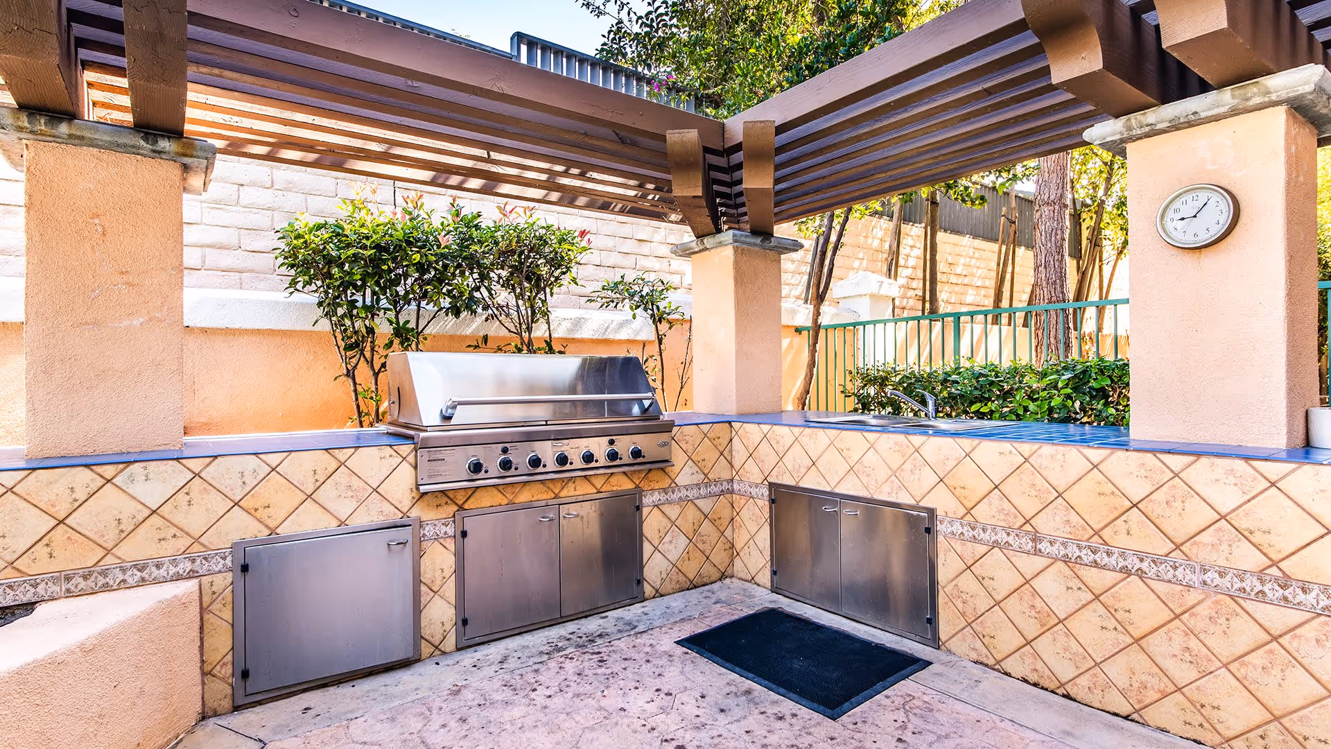 Outdoor grilling area with a stainless steel built-in barbecue grill, tiled countertops, storage cabinets, a sink, and a wall clock mounted on a column under a wooden pergola with greenery and trees in the background.