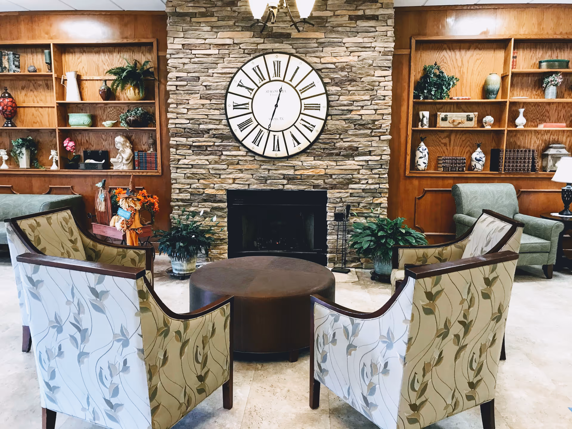 A cozy living room area with four patterned armchairs arranged around a round brown ottoman. Behind the seating is a stone fireplace with a large round clock above it. On either side of the fireplace are wooden built-in shelves decorated with various vases, plants, books, and decorative items. There are also two green upholstered armchairs and some potted plants in the room.