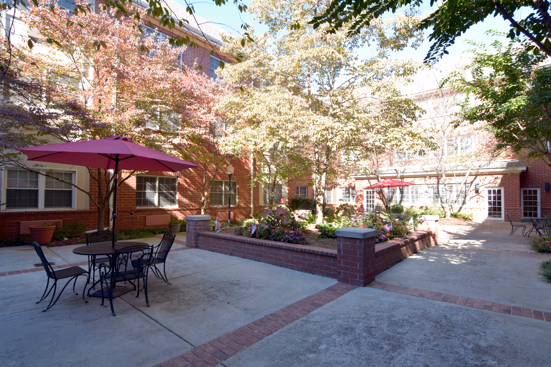 Sunlit brick courtyard with patio tables and red umbrellas, raised flowerbeds, and surrounding multi-story building.