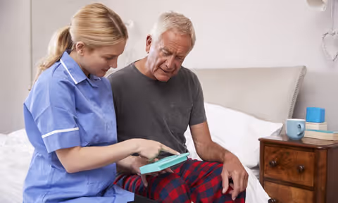 A female caregiver in a blue uniform is sitting on a bed next to an elderly man wearing a gray t-shirt and red plaid pajama pants. They are looking at and pointing to a tablet device together in a bedroom setting with a wooden nightstand holding a blue mug and some books.