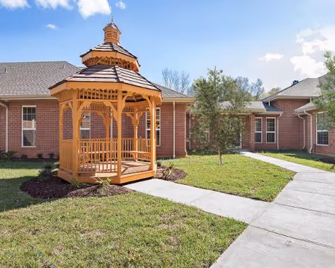Wooden gazebo on a grassy courtyard in front of a red-brick senior living building under a blue sky.