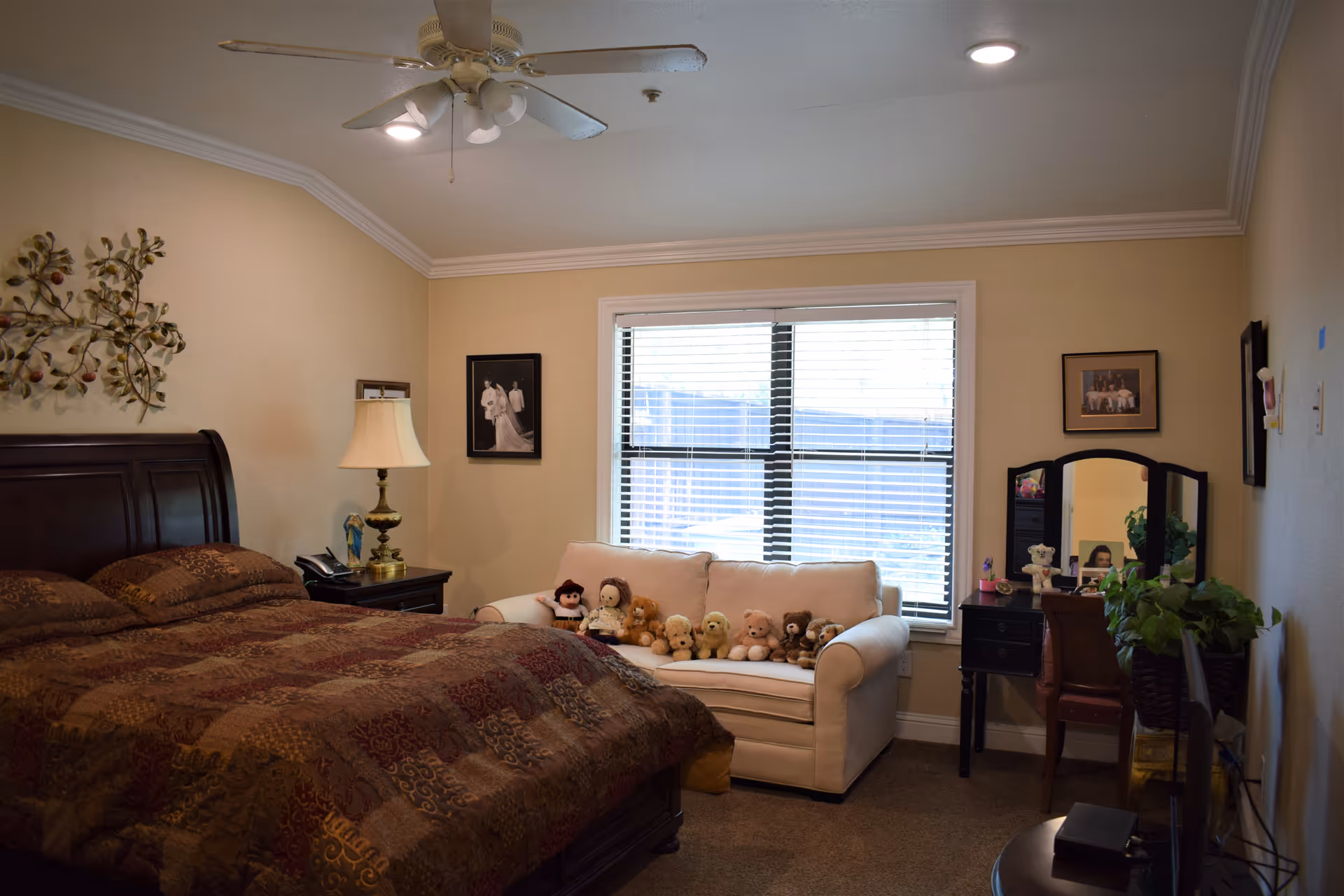 A cozy bedroom with a large dark wooden bed covered in a patterned brown quilt. Next to the bed is a nightstand with a lamp and a telephone. A white loveseat in front of a window with blinds holds several stuffed animals. On the right side of the room is a vanity table with a mirror, a chair, and some plants. The walls are decorated with framed photos and a metal wall art piece. The ceiling has a white fan with lights.
