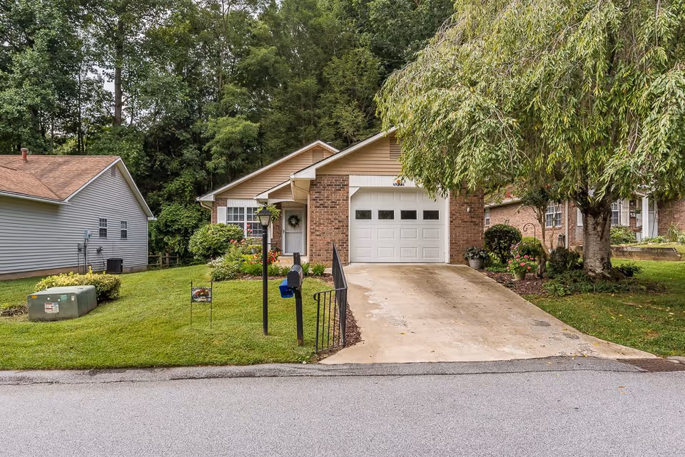A single-story brick house with a white garage door and a driveway. The house is surrounded by a well-maintained lawn with shrubs and flowers. There is a large tree on the right side of the driveway and a mailbox with a blue flag near the street. Another house is visible to the left, and tall trees form the background.