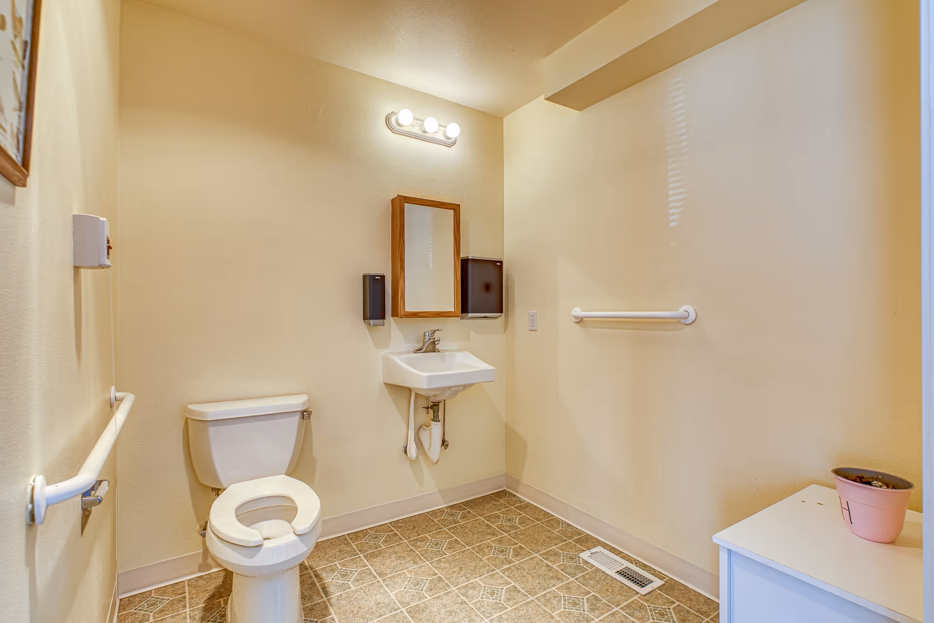A clean, accessible bathroom with a toilet, a wall-mounted sink with a mirror above it, two soap dispensers on either side of the mirror, grab bars on the walls, a tiled floor, and a small white cabinet with a pink potted plant on top.