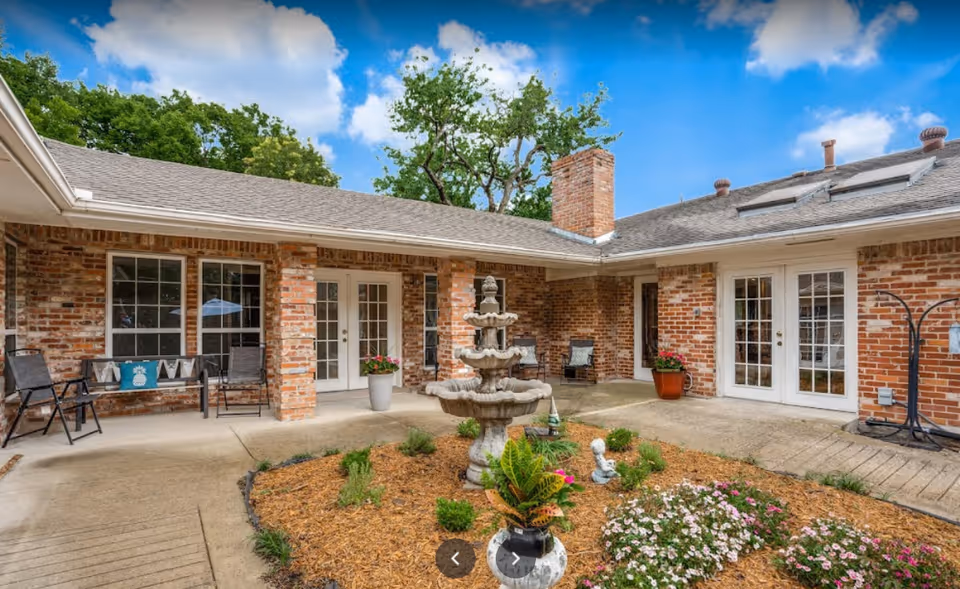 Outdoor courtyard area of a brick building with a three-tiered stone fountain in the center surrounded by mulch and flowers. There are several potted plants, patio chairs, and benches along the walls. The sky is blue with some clouds.
