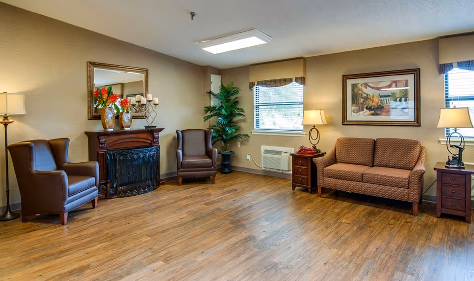 A cozy living room area with two brown leather armchairs, a brown patterned sofa, two wooden side tables with lamps, a decorative fireplace with vases and candles, a large mirror above the fireplace, a potted plant in the corner, two windows with blinds, and a framed painting on the wall.