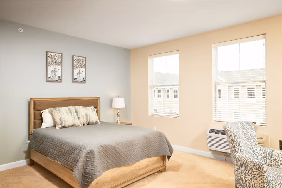 A bright bedroom with a wooden bed frame, a gray quilted bedspread, and three decorative pillows. Two framed botanical prints hang on the light blue wall above the bed. There is a bedside table with a lamp, two large windows with white blinds, a patterned armchair, and a wall-mounted air conditioning unit beneath one window.