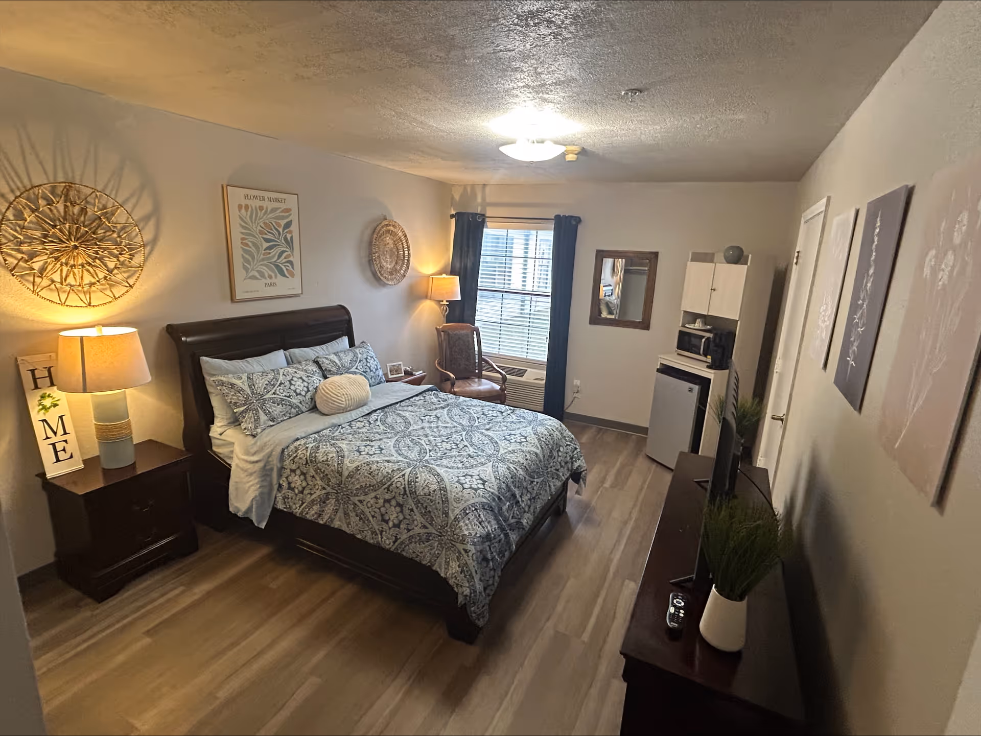 A cozy senior living bedroom with a neatly made bed featuring patterned bedding, a wooden nightstand with a lamp and a decorative 'HOME' sign, a chair near a window with dark curtains, a small kitchenette area with a microwave and mini fridge, a dresser with a TV and potted plants, and wall decorations including framed art and woven baskets.