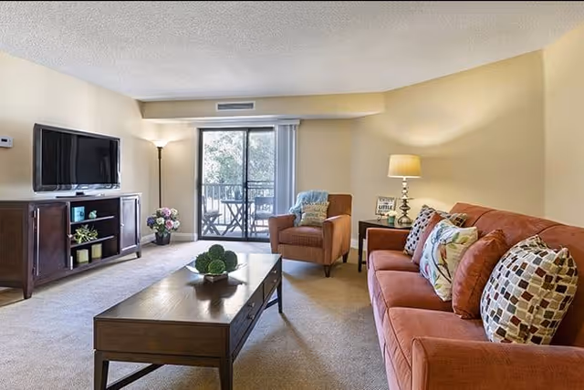 A cozy living room with a large orange sofa adorned with patterned pillows, a wooden coffee table with a decorative centerpiece, a single armchair with a throw blanket, a floor lamp, and a TV mounted on a wooden cabinet. Sliding glass doors lead to a balcony with outdoor seating and greenery visible outside.