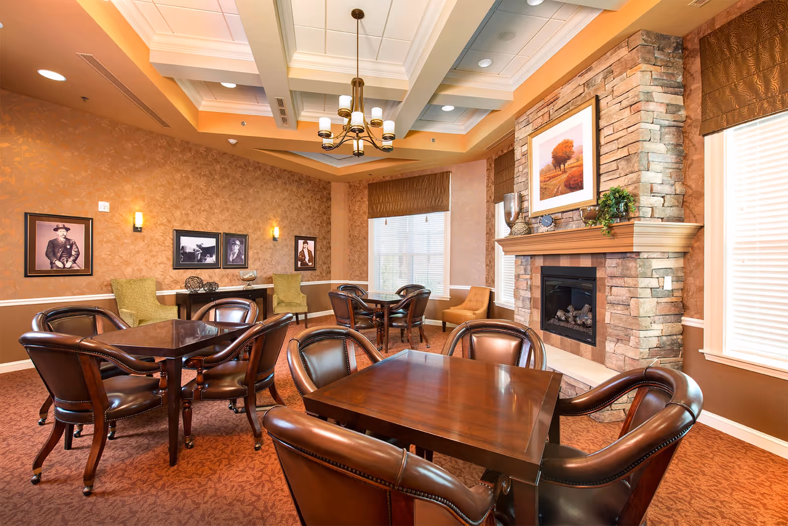 A cozy common area with several wooden tables and leather chairs arranged around them. The room features a stone fireplace with a framed landscape painting above it, warm-toned walls with framed black and white photographs, and large windows with blinds letting in natural light. The ceiling has a decorative tray design with recessed lighting and a chandelier.