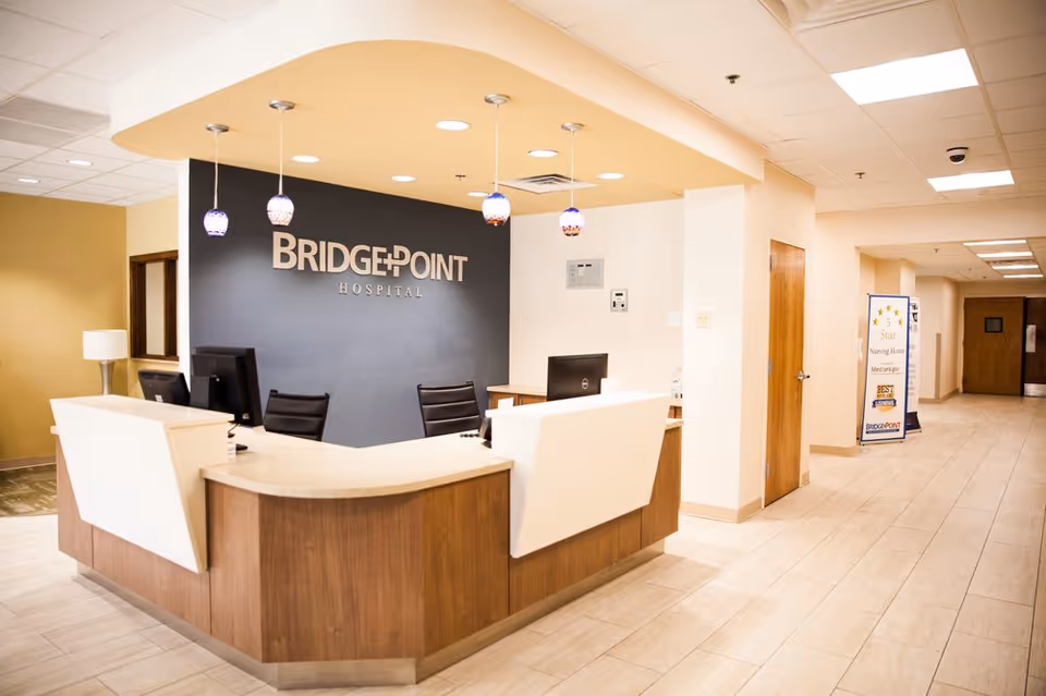 Reception area of BridgePoint Hospital with a modern wooden and white desk, three black chairs, three hanging pendant lights, and a dark blue wall with the BridgePoint Hospital logo. The hallway to the right has doors and a standing sign with awards and recognitions.