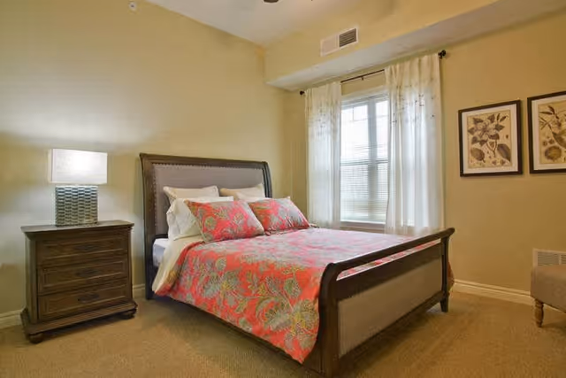 A cozy bedroom with a wooden bed frame featuring a pink floral bedspread and matching pillows. There is a wooden nightstand with a lamp on the left side of the bed. A window with white curtains allows natural light into the room. Two framed botanical prints hang on the wall to the right of the window.