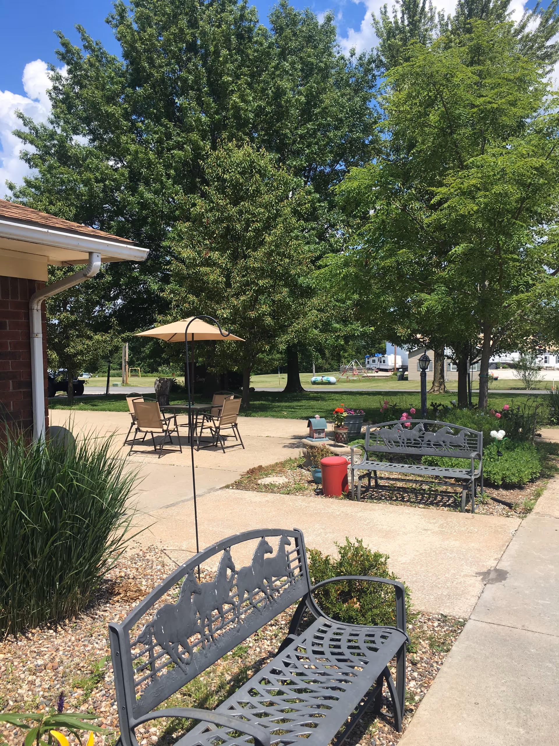 Outdoor patio area at Country View Nursing Facility with metal benches featuring horse designs, a table with chairs under a beige umbrella, green trees, and a clear blue sky.
