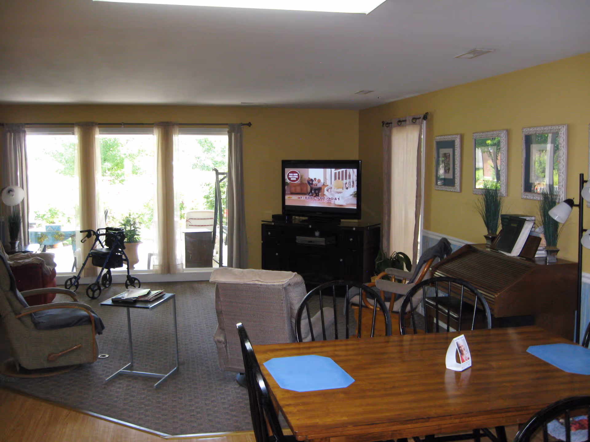 Open living and dining area with a wooden dining table in the foreground, seating and a TV by large windows, and a walker near the sliding doors.
