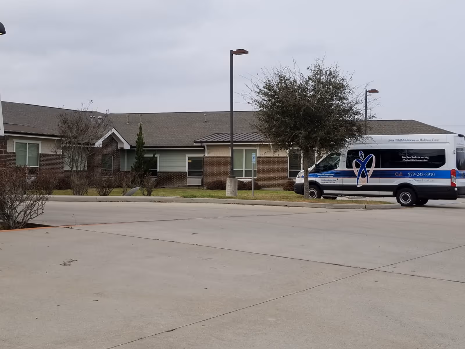 Exterior view of Arbor Hills Rehabilitation and Healthcare Center showing a single-story brick and siding building with a parking lot in front. A white and blue shuttle van with the facility's name and contact information is parked near a tree and a streetlamp.