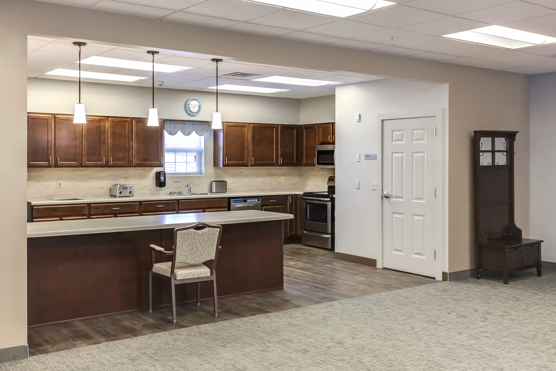 A spacious kitchen area in an assisted living facility featuring dark wood cabinets, a long countertop with a single chair, stainless steel appliances including a stove and microwave, a toaster, and a small window with a clock above it. The floor is a combination of wood and carpet, and there is a wooden hall tree with hooks and a bench against the wall near a closed white door.