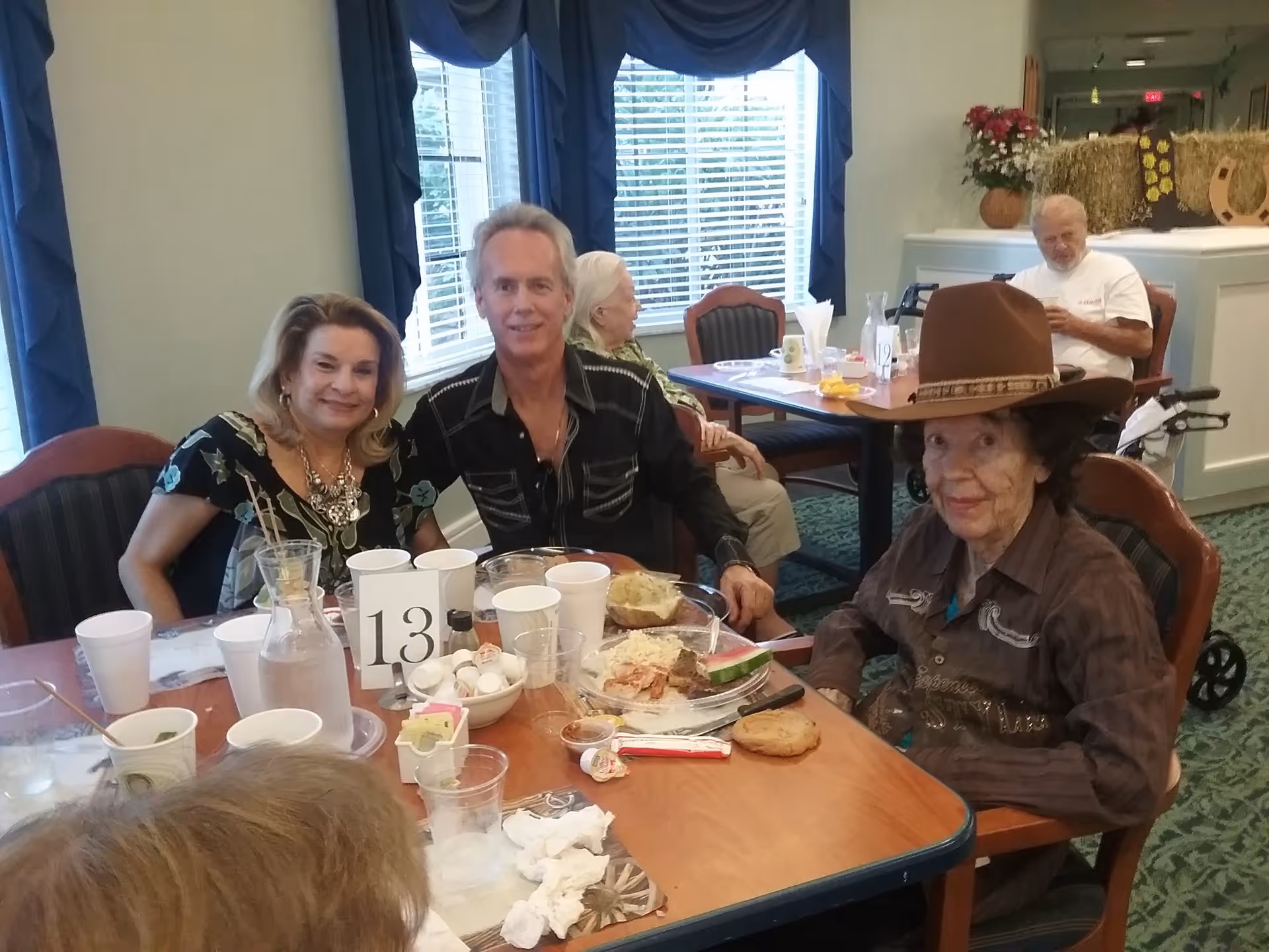 A group of elderly people and a middle-aged man sitting around a dining table in a senior living facility. The table has plates with food, cups, and a table number 13. One elderly woman is wearing a large brown cowboy hat. The room has large windows with blue curtains and a green carpet.