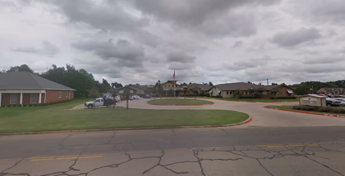 Exterior view of Spring Lake Assisted Living and Memory Care facility showing a circular driveway with a flagpole in the center, multiple single-story buildings, parked vehicles, and a sign with the facility's name on the right side. The sky is cloudy.
